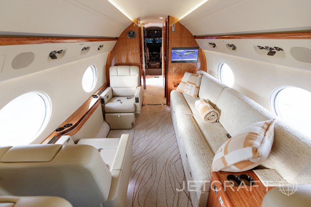 A wide view of the business jet's luxurious cabin, showing a cream sofa, individual seats, and a wall-mounted screen.
